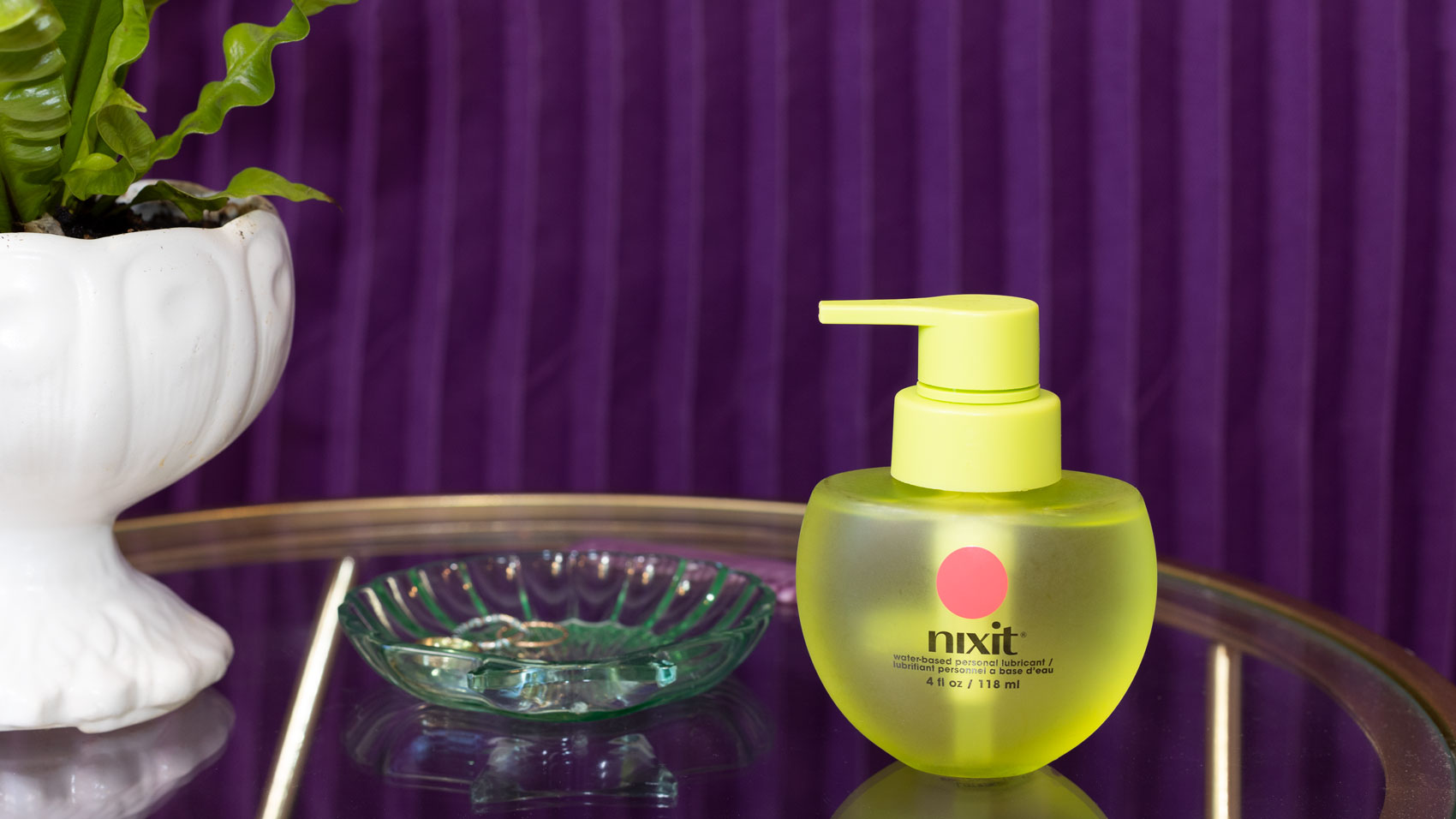 A round yellow nixit foaming cleanser bottle sits next to a clear glass dish on a mirrored surface, with a potted plant and a purple striped background.
