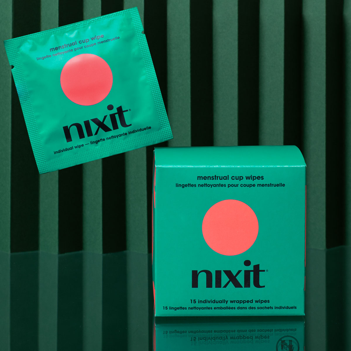 A nixit menstrual cup wipe packet is displayed above a matching box of 15 wipes, both teal with a large pink circle, against a green striped background.