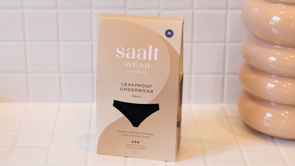 A box of Saalt Wear leakproof bikini underwear in medium size sits on a white tiled surface next to a beige ceramic vase.