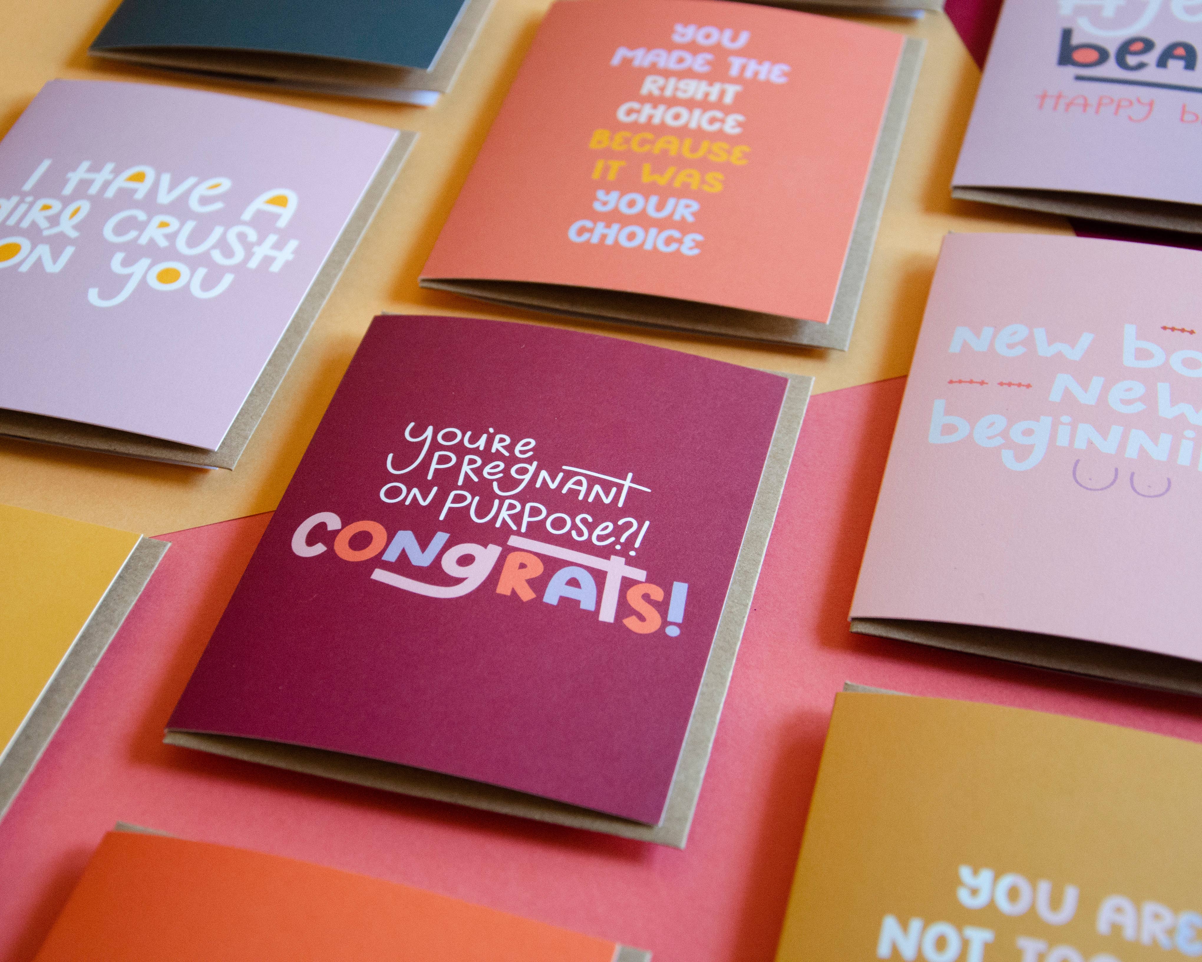 The "You're Pregnant on Purpose Card" is featured among several colorful greeting cards arranged on a pink and orange surface, offering celebratory and supportive messages—perfect as pregnancy congratulations cards.