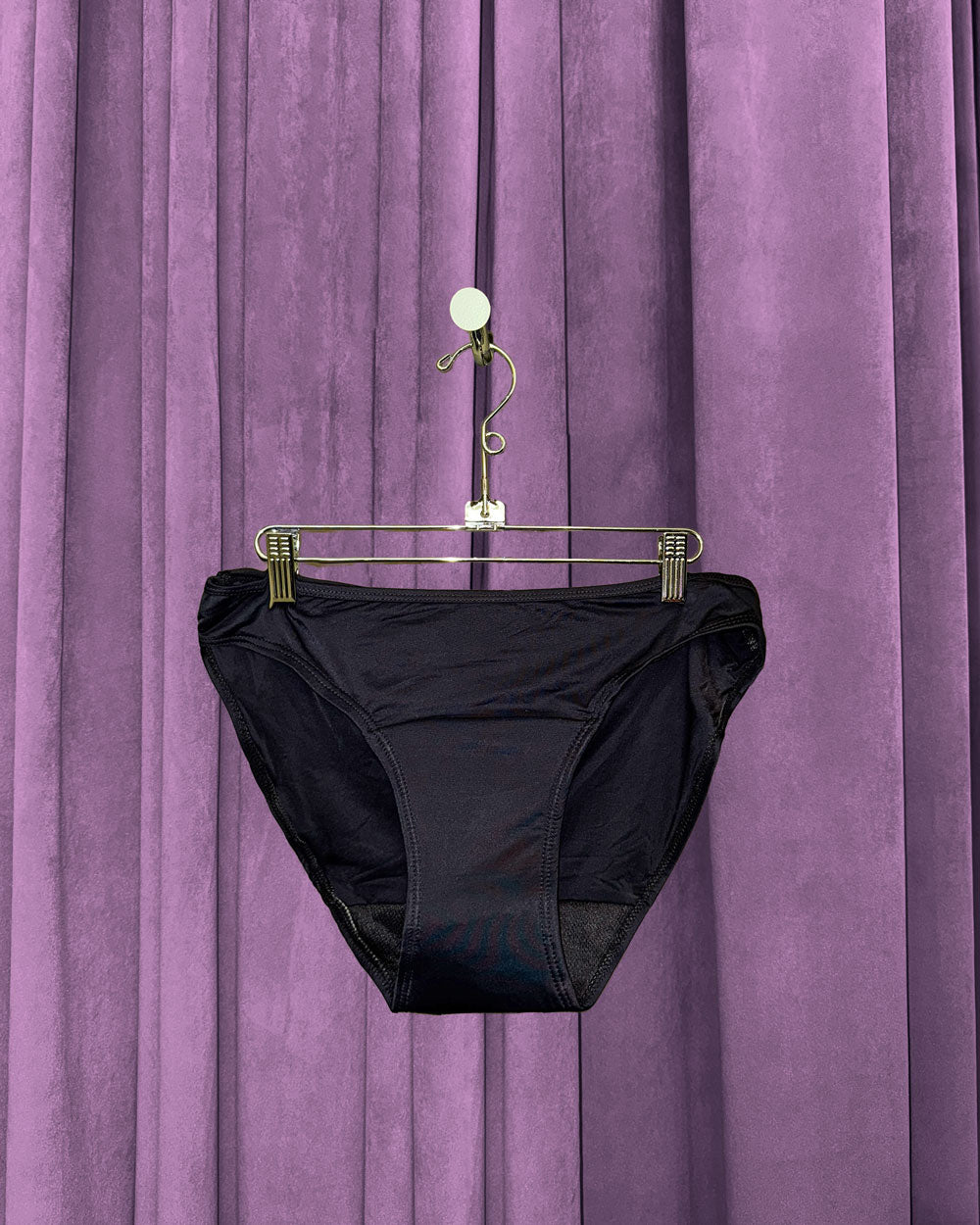 A pair of Period Swim Bottoms - Bikini Cut in black hangs on a metal hanger in front of purple velvet curtains.