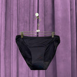 A pair of Period Swim Bottoms - Bikini Cut in black hangs on a metal hanger in front of purple velvet curtains.