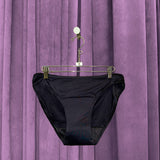 A pair of Period Swim Bottoms - Bikini Cut in black hangs on a metal hanger in front of purple velvet curtains.