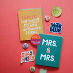Two colorful greeting cards on a red background—one reading "Thanks for being my bad influence friend," the other "MRS. & MRS."—are surrounded by pins and a My Body My Choice Sticker, plus stickers with phrases like Vote Blue and Stay Angry.