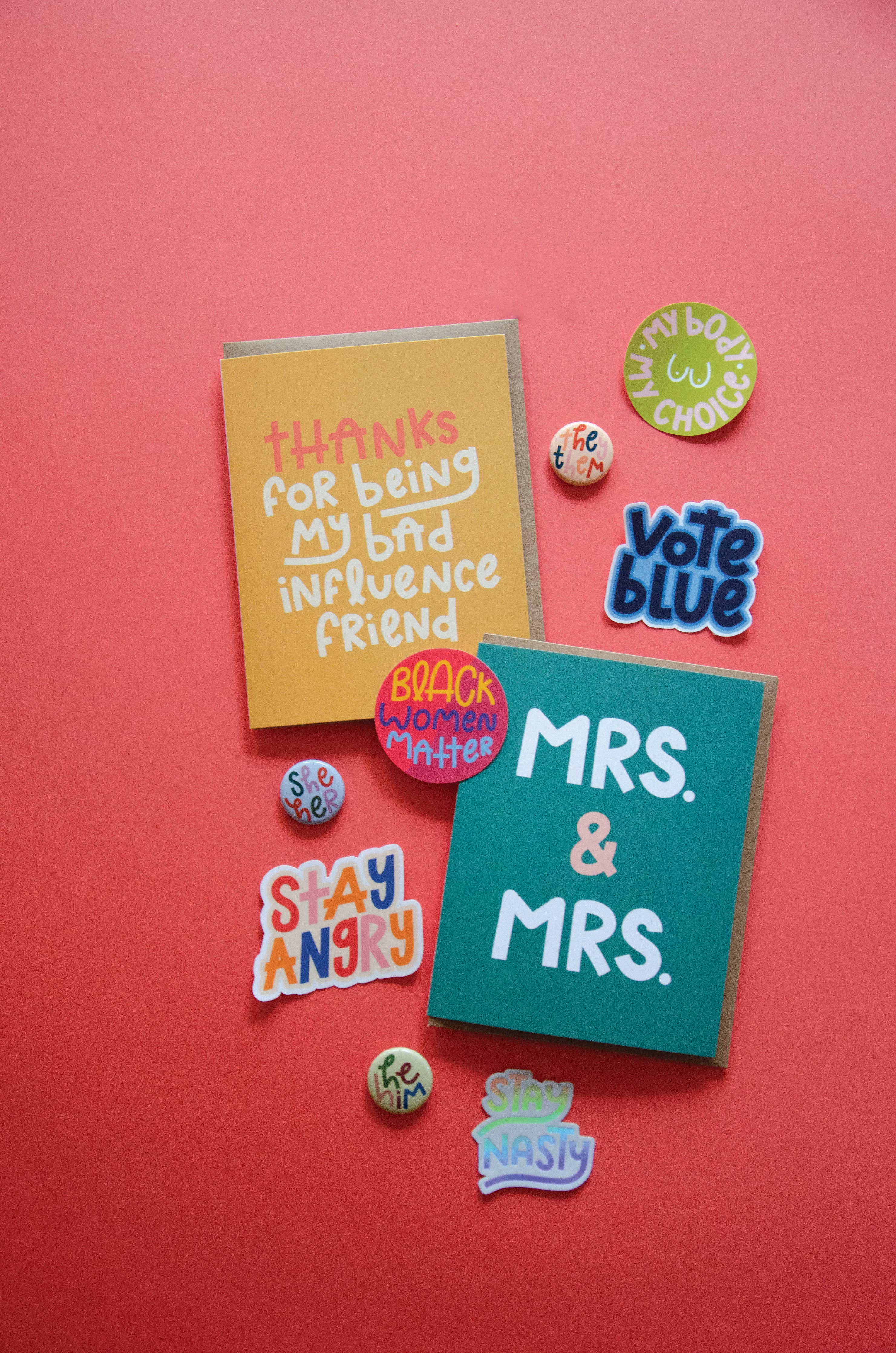 Two colorful greeting cards on a red background—one reading "Thanks for being my bad influence friend," the other "MRS. & MRS."—are surrounded by pins and a My Body My Choice Sticker, plus stickers with phrases like Vote Blue and Stay Angry.