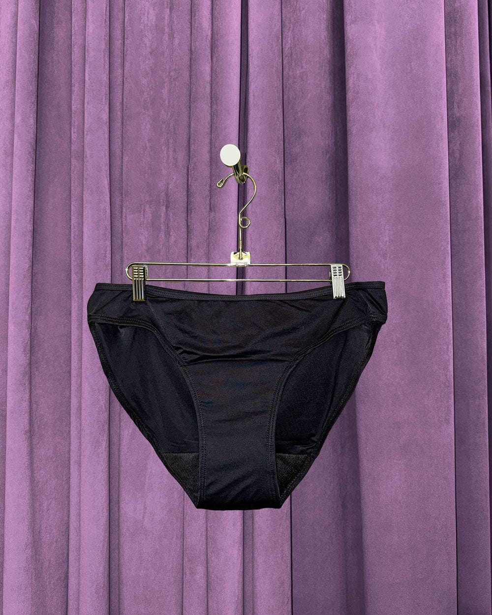 A pair of Period Swim Bottoms - Bikini Cut hangs on a hanger with clips in front of a purple velvet curtain.