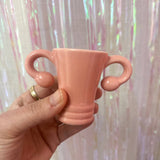 A hand holds the Shooterus - Uterus Shot Glass, a small pink ceramic cup shaped like a uterus with fallopian tube handles, perfect as a novelty item or conversation starter about menopause. Displayed against an iridescent curtain background.
