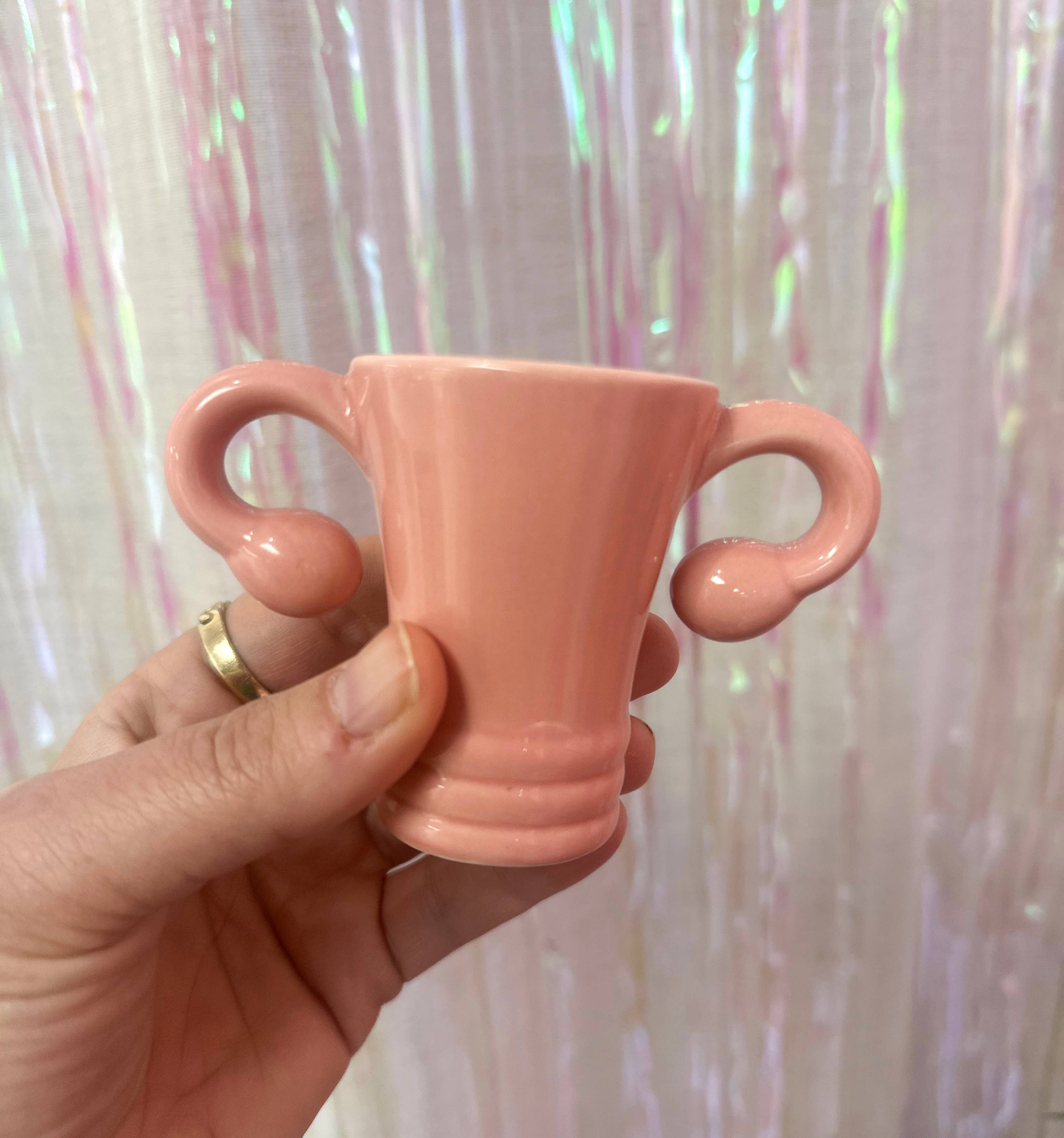 A hand holds the Shooterus - Uterus Shot Glass, a small pink ceramic cup shaped like a uterus with fallopian tube handles, perfect as a novelty item or conversation starter about menopause. Displayed against an iridescent curtain background.