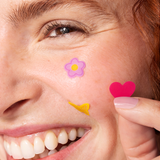 A close-up of a smiling person’s face with a pink flower and yellow butterfly sticker on their cheek, plus Big Hug Breakout Pimple Patches. They’re holding a pink heart-shaped sticker near their cheek with their fingers.