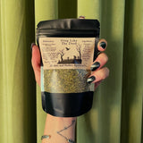 A hand with black, glittery nails holds a Sleep Like The Dead Tea by Haints and Hollers Apothecary packet—an herbal blend with valerian root. The label lists ingredients, instructions, and the dried herbs show through in front of green curtains.