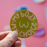 A hand holds the My Body My Choice Sticker, a round green women's rights sticker featuring “MY BODY MY CHOICE” encircling a line-drawn torso, shown against a pink background.