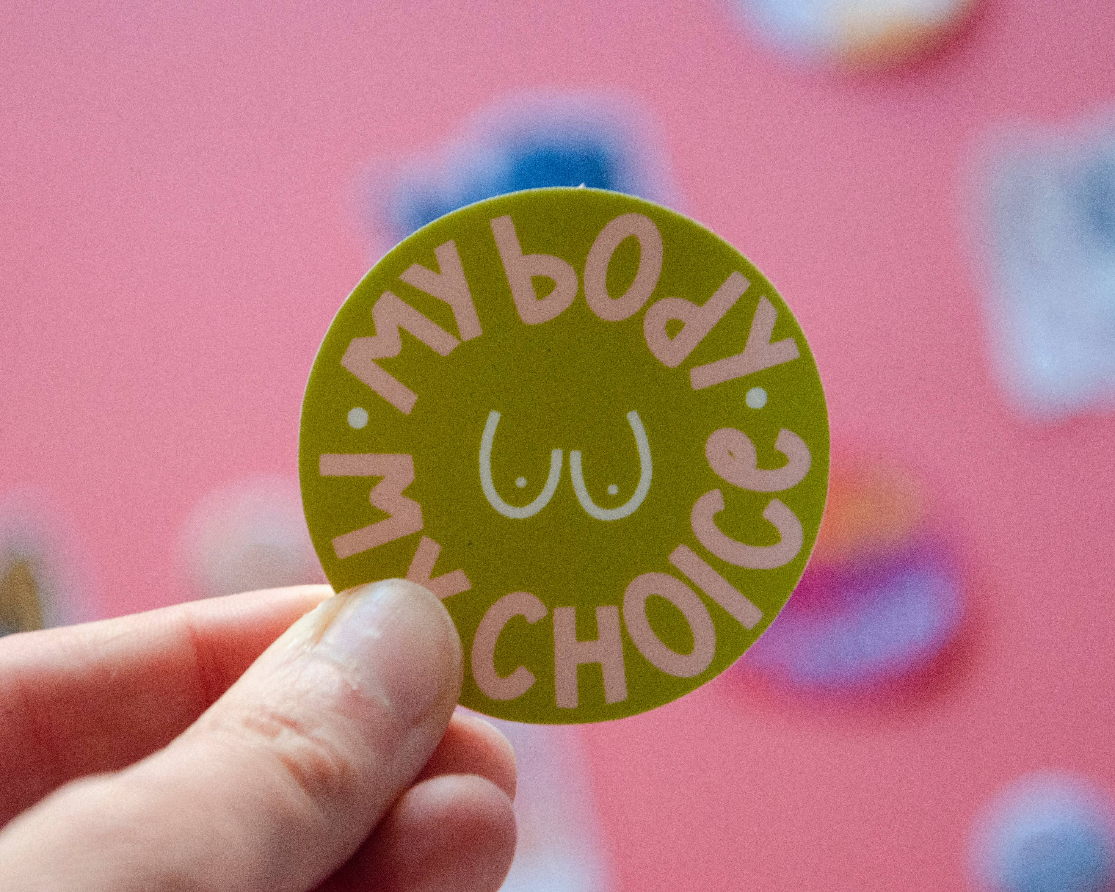 A hand holds the My Body My Choice Sticker, a round green women's rights sticker featuring “MY BODY MY CHOICE” encircling a line-drawn torso, shown against a pink background.