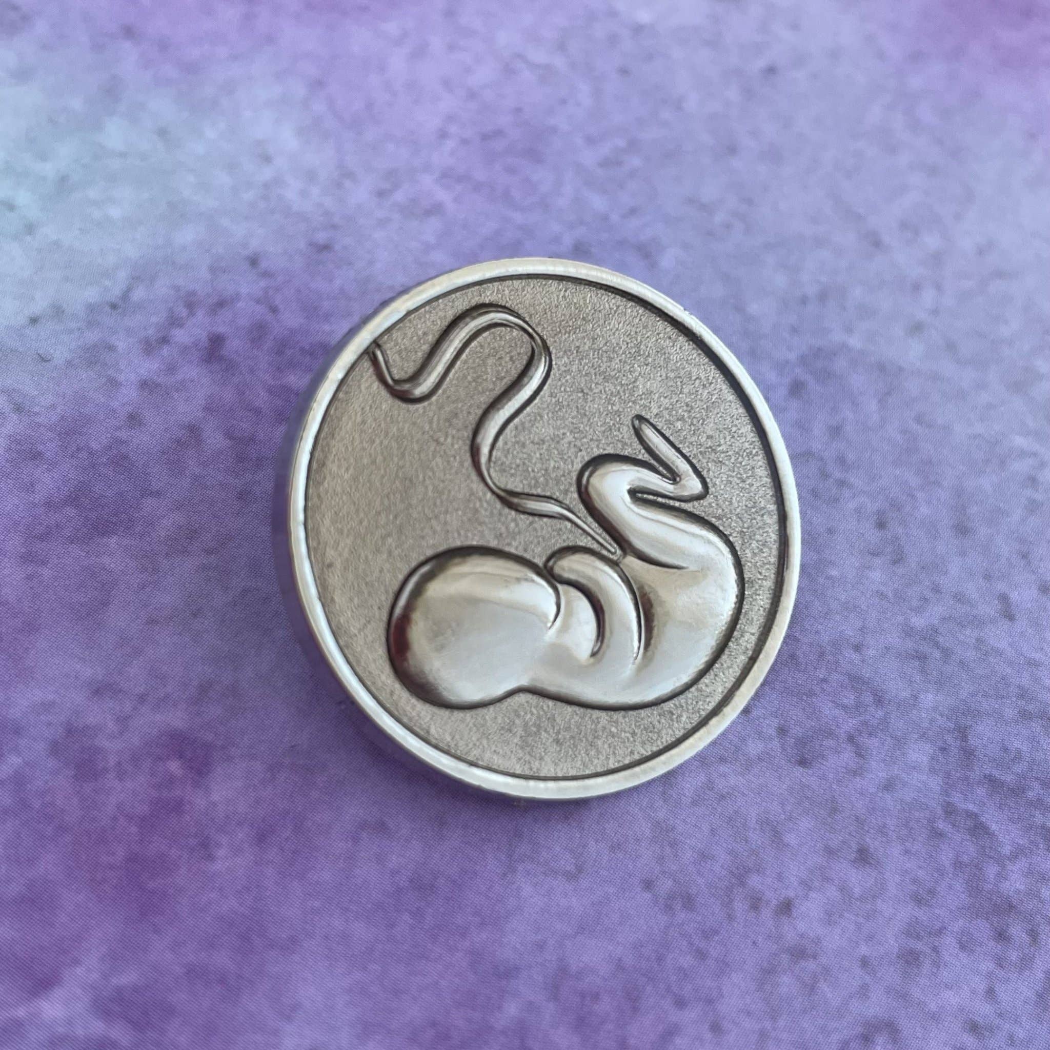 The OB/GYN Pin Pack includes a round silver uterus pin with a raised fetus and umbilical cord on a textured purple background—an elegant anatomy accessory ideal for OB GYN professionals or enthusiasts.