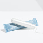 A Lola 20ct Regular Tampon made of white organic cotton with a BPA-free applicator is shown in front of its blue LOLA wrapper, set on a plain white surface with a light gray background.