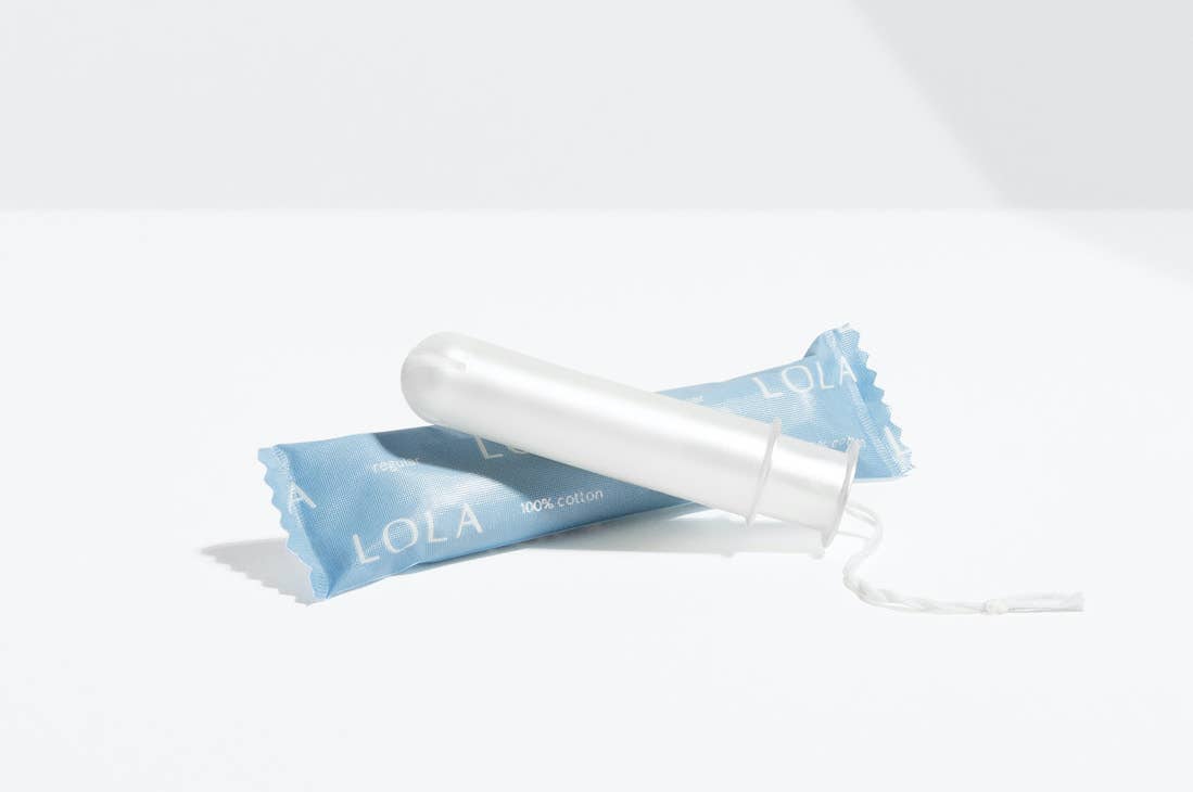 A Lola 20ct Regular Tampon made of white organic cotton with a BPA-free applicator is shown in front of its blue LOLA wrapper, set on a plain white surface with a light gray background.