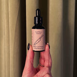 A hand with black nail polish holds a dark glass dropper bottle labeled Red Moon Tincture by Haints and Hollers Apothecary, designed for menstrual comfort. A knife illustration appears on the label, with cream-colored curtains in the background.
