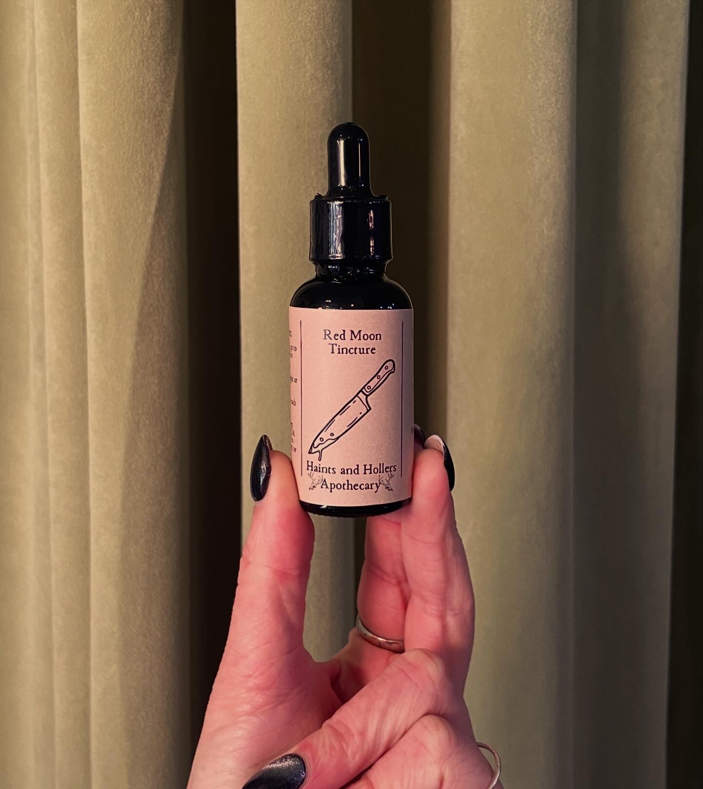 A hand with black nail polish holds a dark glass dropper bottle labeled Red Moon Tincture by Haints and Hollers Apothecary, designed for menstrual comfort. A knife illustration appears on the label, with cream-colored curtains in the background.
