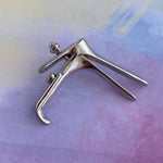 An elegant uterus pin from the OB/GYN Pin Pack sits beside a metal speculum on a pastel surface in shades of purple, pink, and yellow.