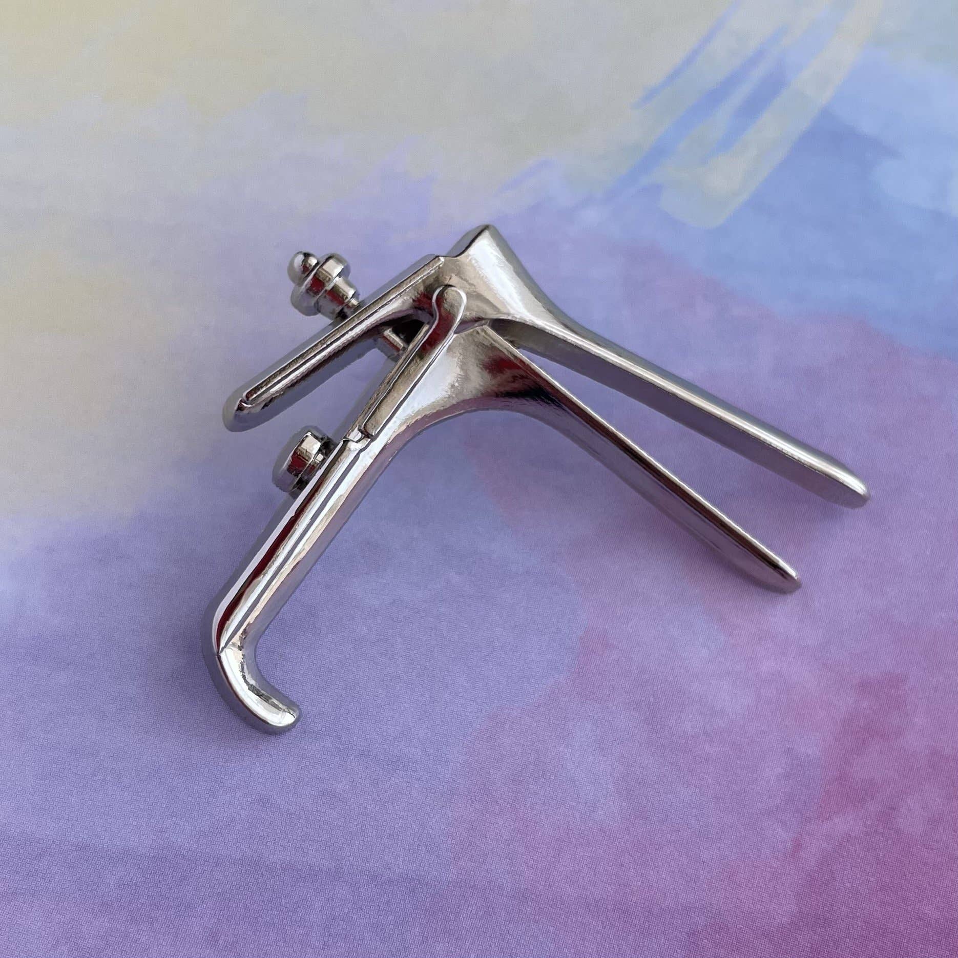An elegant uterus pin from the OB/GYN Pin Pack sits beside a metal speculum on a pastel surface in shades of purple, pink, and yellow.