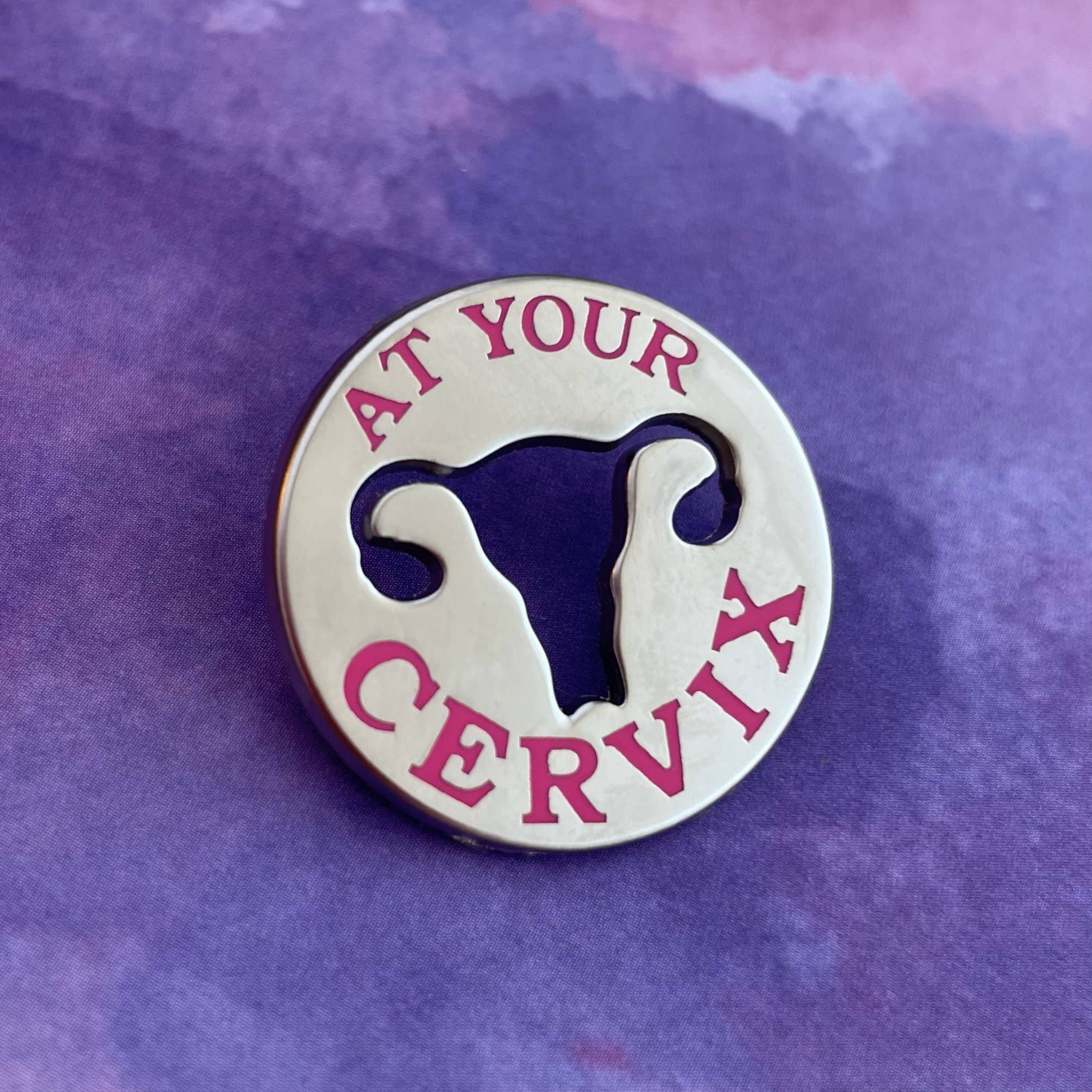 The OB/GYN Pin Pack features a round metal pin with a uterus-shaped cutout, pink "AT YOUR CERVIX" text on a purple watercolor background—perfect for OB GYN pin collectors.