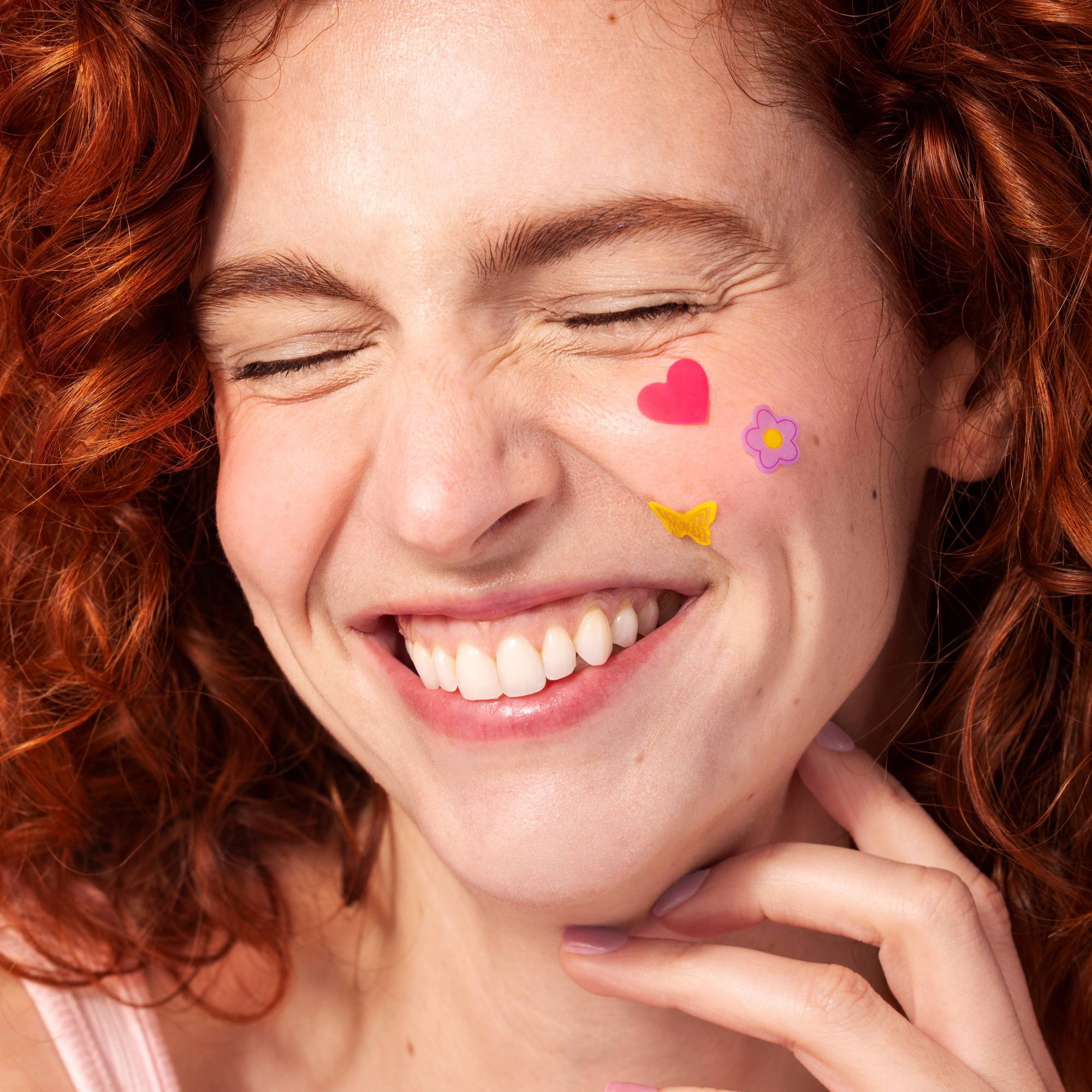 A woman with curly red hair smiles with her eyes closed, touching her face adorned with Big Hug Breakout Pimple Patches—colorful hydrocolloid patches shaped like a pink heart, yellow butterfly, and purple flower on her cheek.