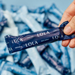 A hand holds a dark blue Lola 20ct Super Plus Tampons wrapper, with more tampons scattered in the background.