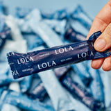 A hand holds a dark blue Lola 20ct Super Plus Tampons wrapper, with more tampons scattered in the background.