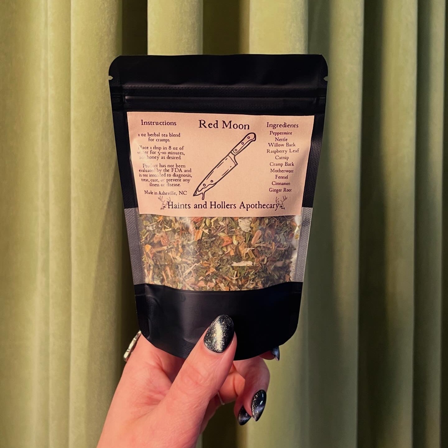 A hand with black, sparkly nail polish holds a packet labeled Red Moon Tea by Haints and Hollers Apothecary in front of green curtains. The packet lists instructions and ingredients for this herbal tea blend made to soothe menstrual cramps.