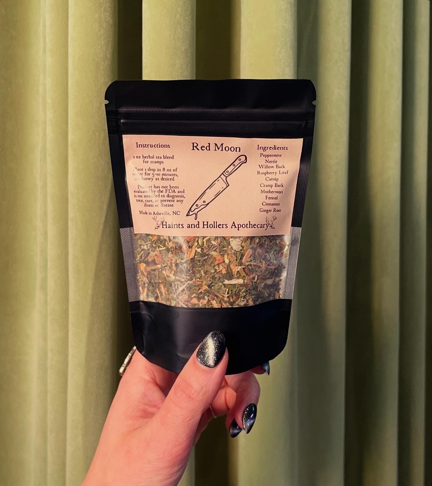A hand with black, sparkly nail polish holds a packet labeled Red Moon Tea by Haints and Hollers Apothecary in front of green curtains. The packet lists instructions and ingredients for this herbal tea blend made to soothe menstrual cramps.