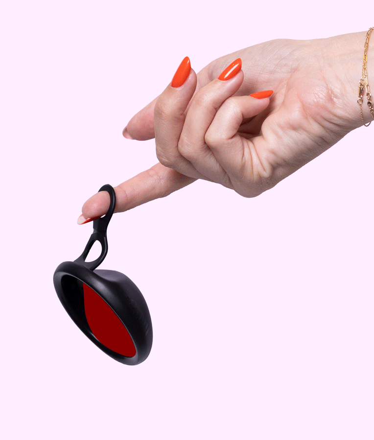 A hand with red manicured nails and a gold bracelet holds the Hello Disc - Black, a circular keychain device with a double loop tab, balanced on the index finger against a pale pink background for easy removal.