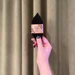 A hand with black nail polish holds up a Pennyroyal Interlabial Pad, wrapped in its label and reminiscent of absorbent cotton pads, against a sage green curtain background.