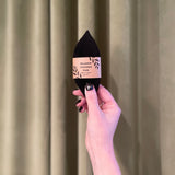 A hand with black nail polish holds up a Pennyroyal Interlabial Pad, wrapped in its label and reminiscent of absorbent cotton pads, against a sage green curtain background.