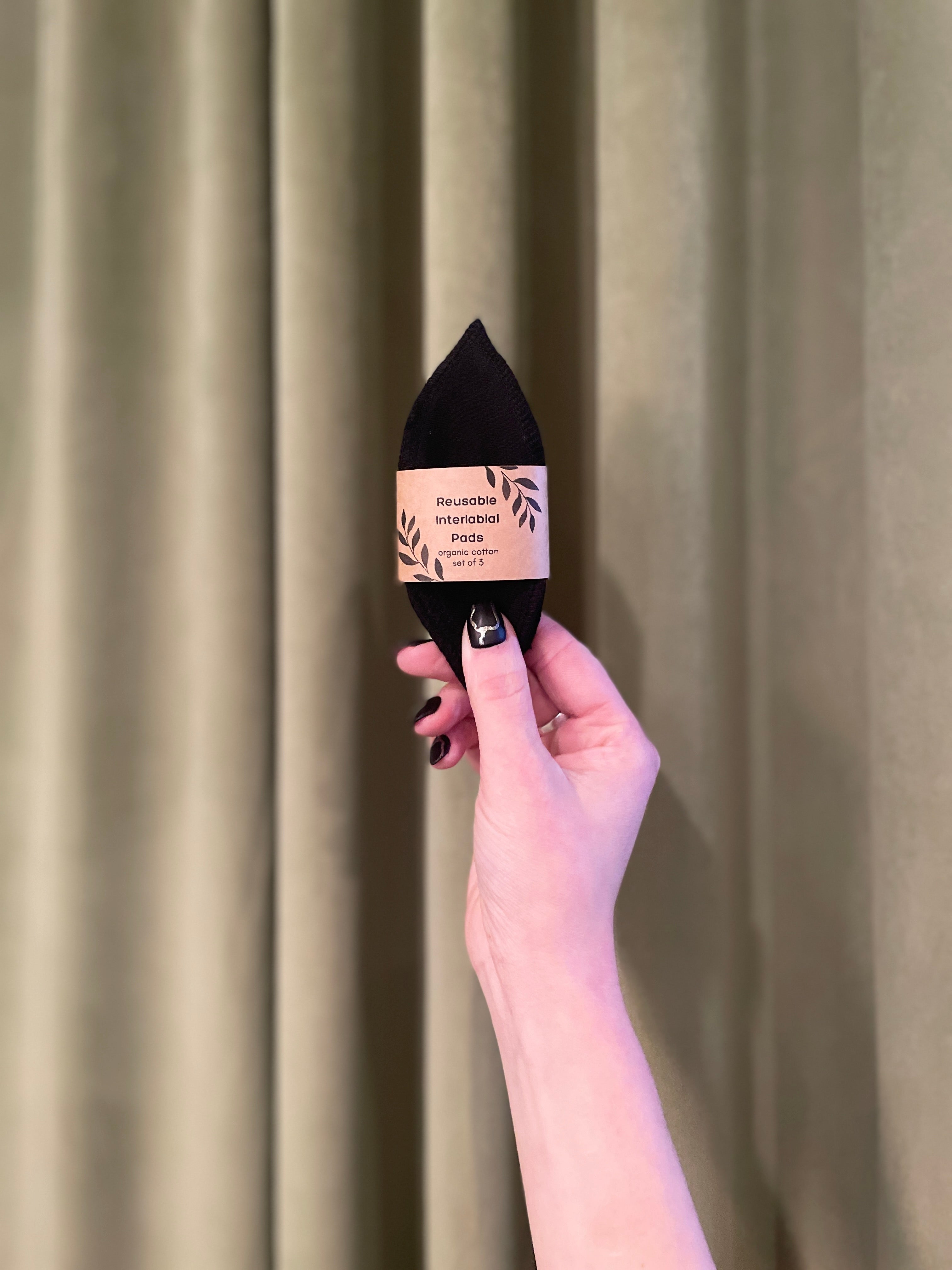 A hand with black nail polish holds up a Pennyroyal Interlabial Pad, wrapped in its label and reminiscent of absorbent cotton pads, against a sage green curtain background.