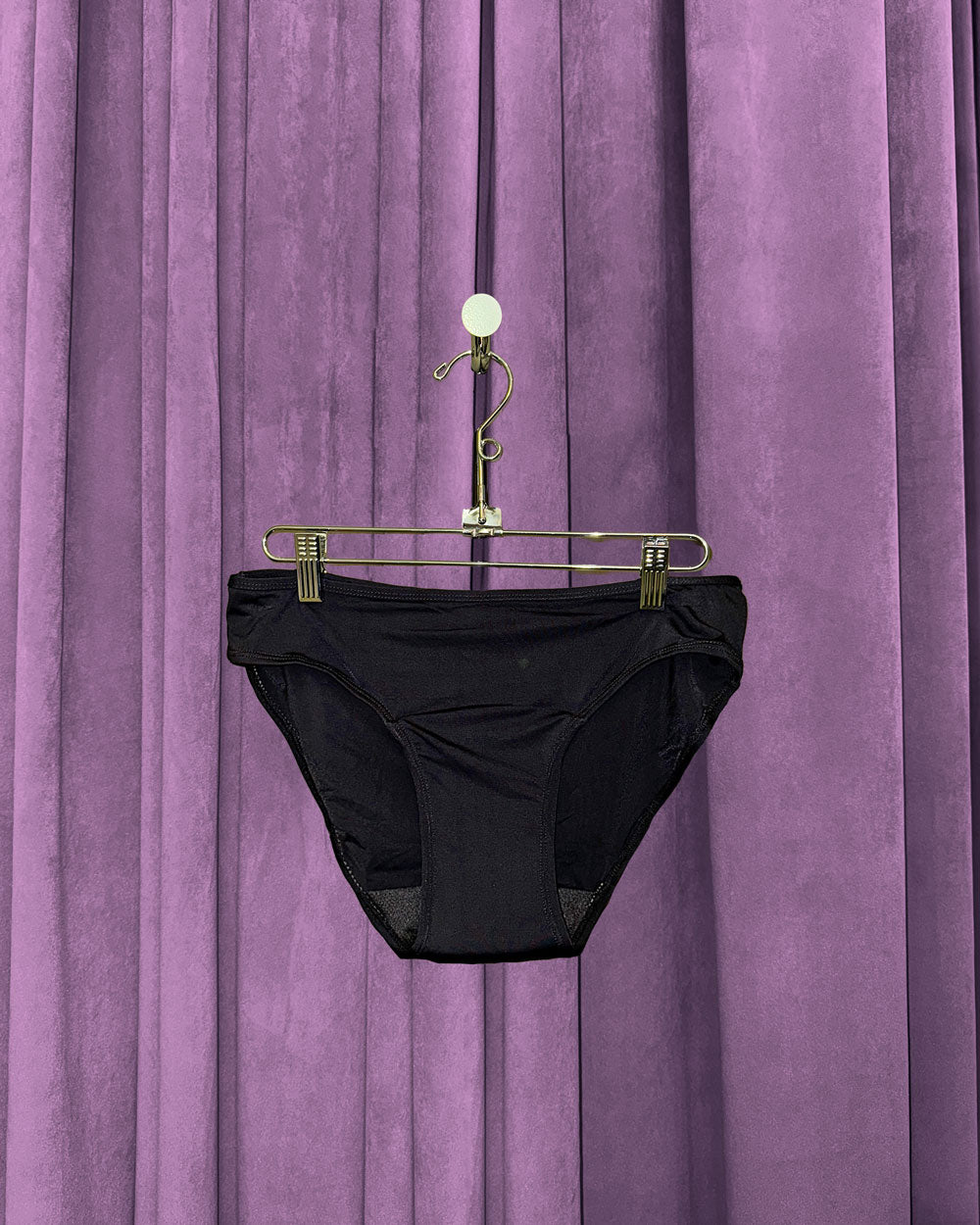 A pair of Period Swim Bottoms - Bikini Cut in black hangs on a gold hanger with clips, set against closed purple curtains.