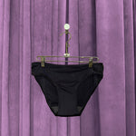 A pair of Period Swim Bottoms - Bikini Cut in black hangs on a gold hanger with clips, set against closed purple curtains.