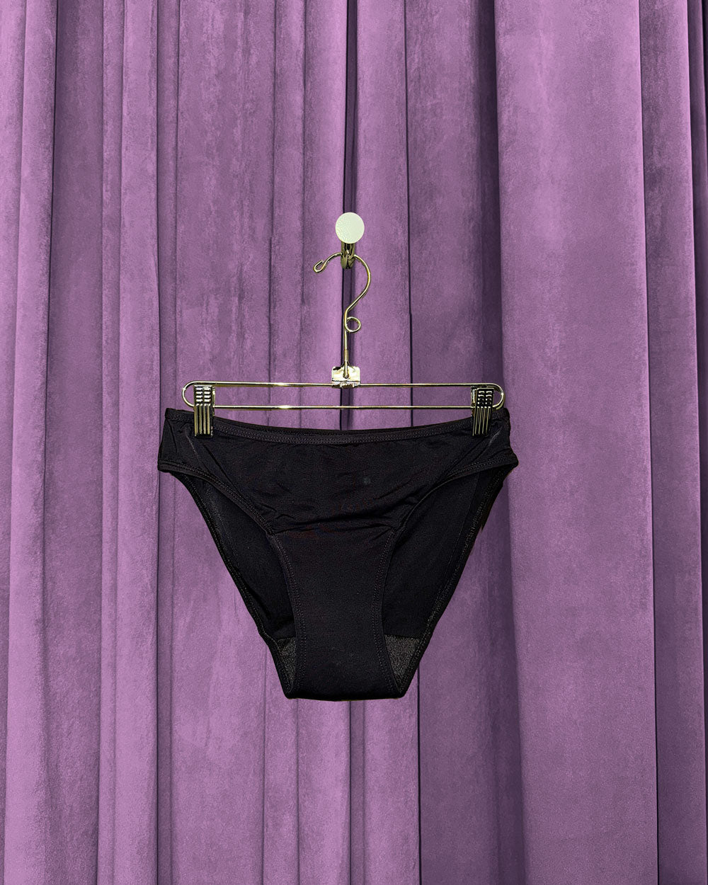 A pair of Period Swim Bottoms - Bikini Cut in black hangs on a metal hanger in front of closed purple velvet curtains.