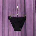 A pair of Period Swim Bottoms - Bikini Cut in black hangs on a metal hanger in front of closed purple velvet curtains.