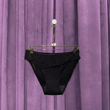 A pair of Period Swim Bottoms - Bikini Cut in black hangs on a metal hanger in front of closed purple velvet curtains.