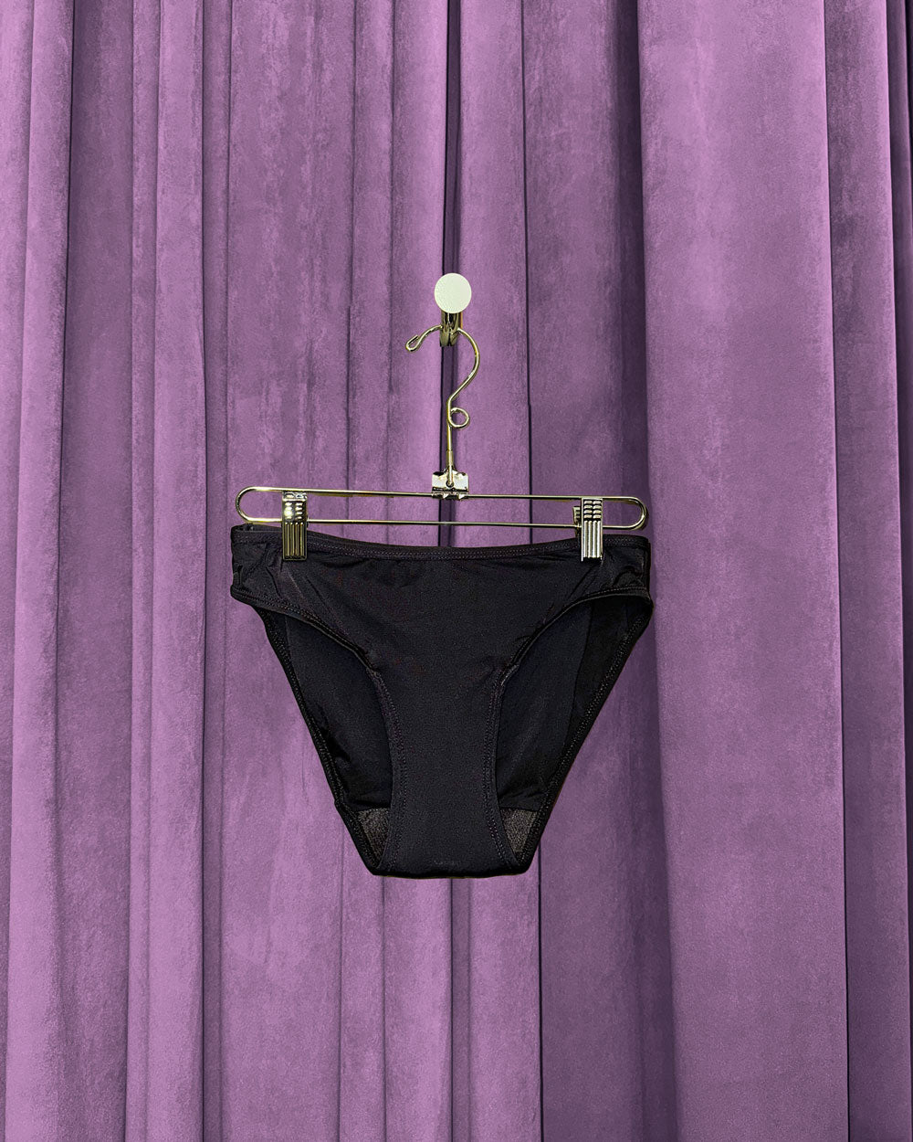 A pair of Period Swim Bottoms - Bikini Cut in black hangs on a gold hanger in front of closed, purple velvet curtains.