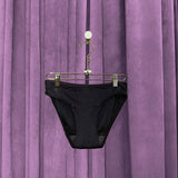 A pair of Period Swim Bottoms - Bikini Cut in black hangs on a gold hanger in front of closed, purple velvet curtains.