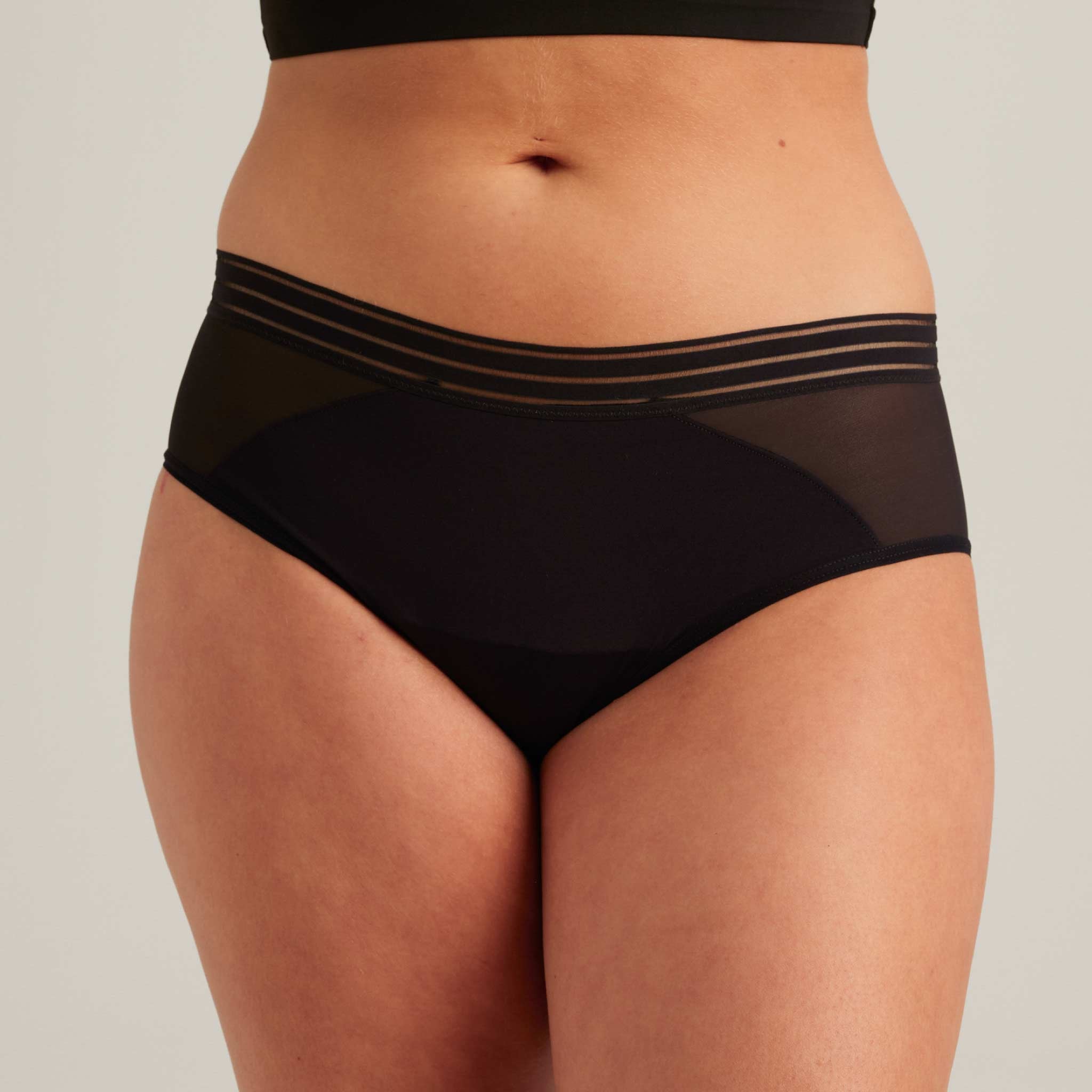 A person wears the Saalt Wear Leakproof Mesh Hipster—black underwear with sheer paneling and a striped waistband—shown from mid-torso to upper thighs against a neutral background.