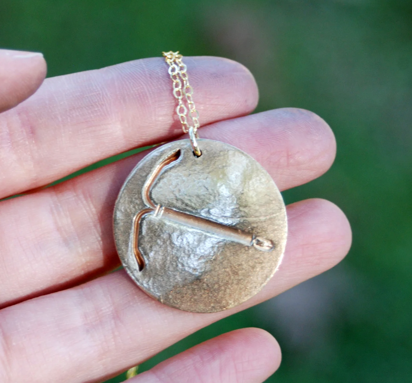 A hand holds the IUD Necklace, which is round and bronze with an embossed pickaxe design on a gold chain. The blurred green background creates an outdoor atmosphere.