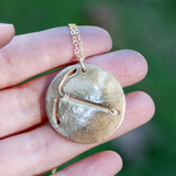 A hand holds the IUD Necklace, which is round and bronze with an embossed pickaxe design on a gold chain. The blurred green background creates an outdoor atmosphere.