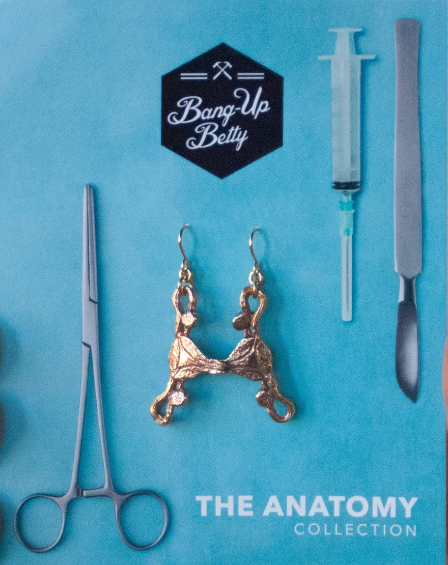 Uterus Earrings: Gold, pelvic bone-shaped earrings shown on blue with surgical tool illustrations, a syringe, and "Bang-Up Betty THE ANATOMY COLLECTION"—ideal for fans of unique uterus jewelry.