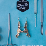Uterus Earrings: Gold, pelvic bone-shaped earrings shown on blue with surgical tool illustrations, a syringe, and "Bang-Up Betty THE ANATOMY COLLECTION"—ideal for fans of unique uterus jewelry.