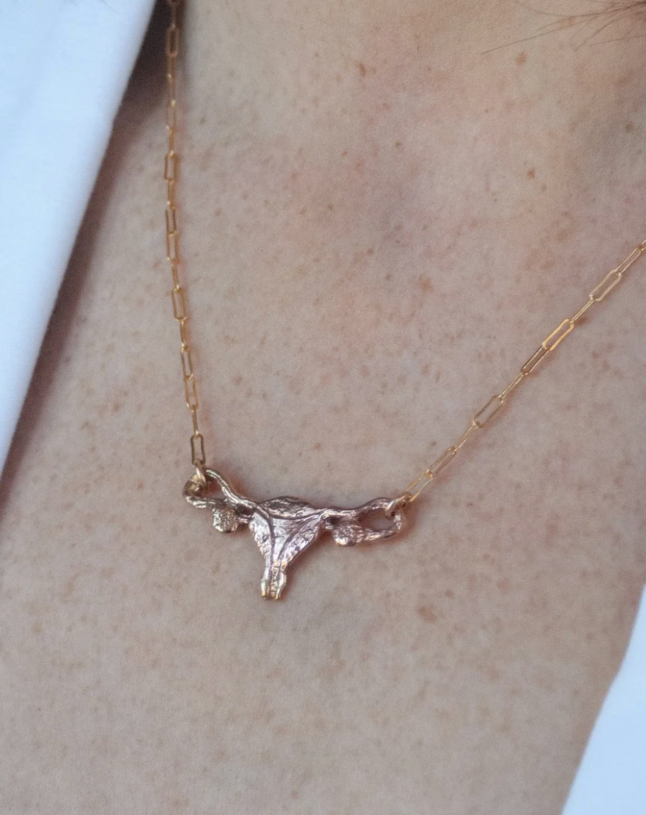A close-up of a person wearing the Bronze Uterus Necklace by Bang-Up Betty—a striking bronze pendant on a chain, shown against light skin and a white garment from the creator known for bold statement jewelry.