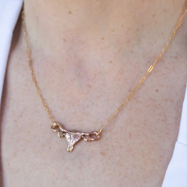 A Sterling Silver Uterus Necklace by Bang-Up Betty, featuring a delicate uterus-shaped pendant worn around the neck against light skin and a white shirt.