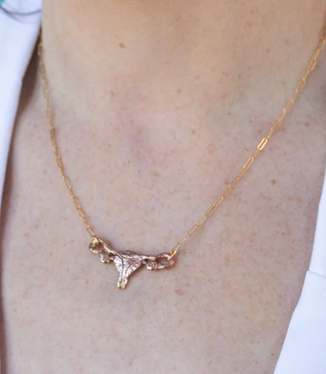 A Sterling Silver Uterus Necklace by Bang-Up Betty, featuring a delicate uterus-shaped pendant worn around the neck against light skin and a white shirt.