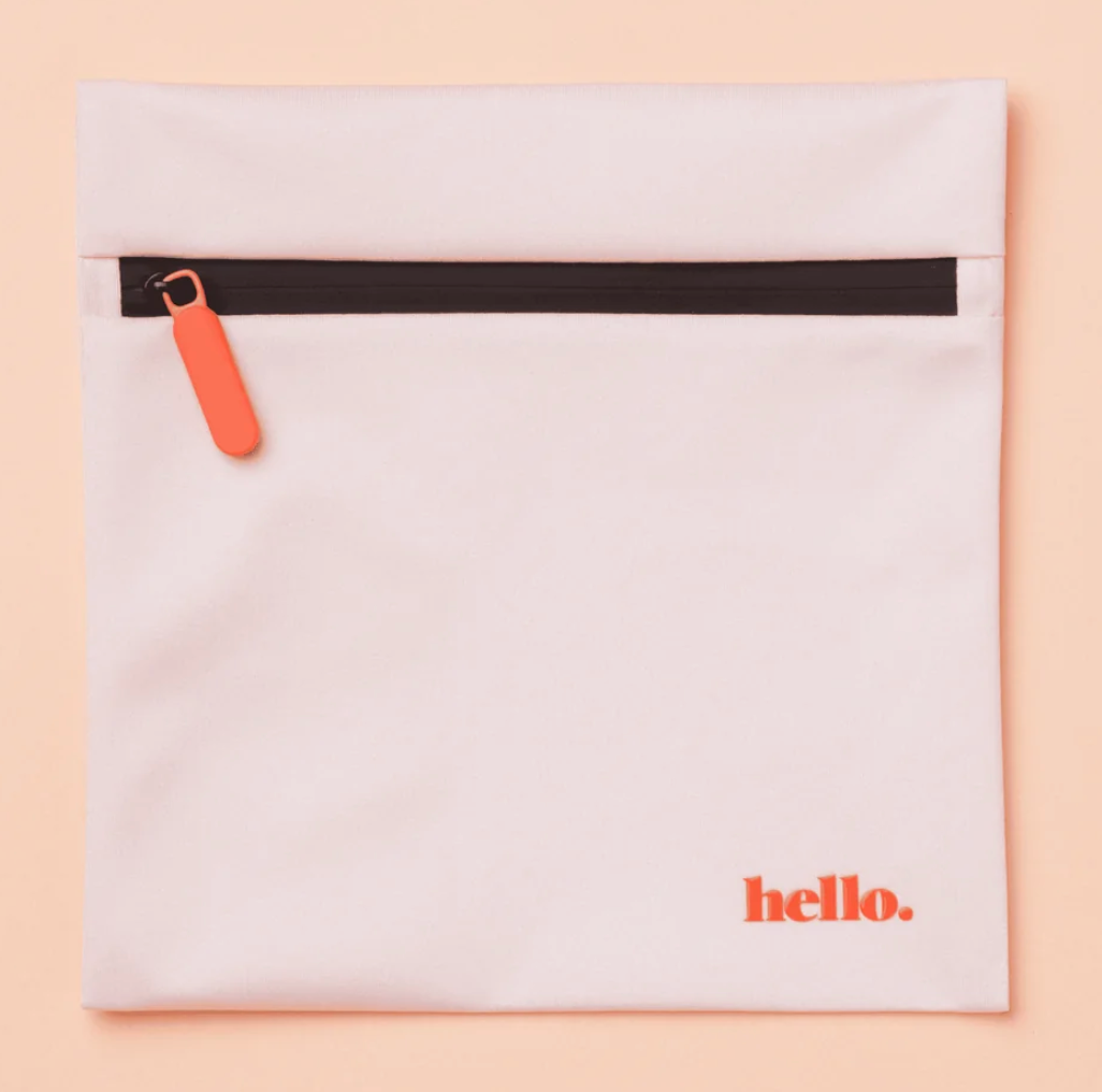 The Hello Wet Bag is a light pink waterproof storage bag featuring an orange zipper pull and "hello." printed in orange in the lower right corner, shown against a soft peach background.
