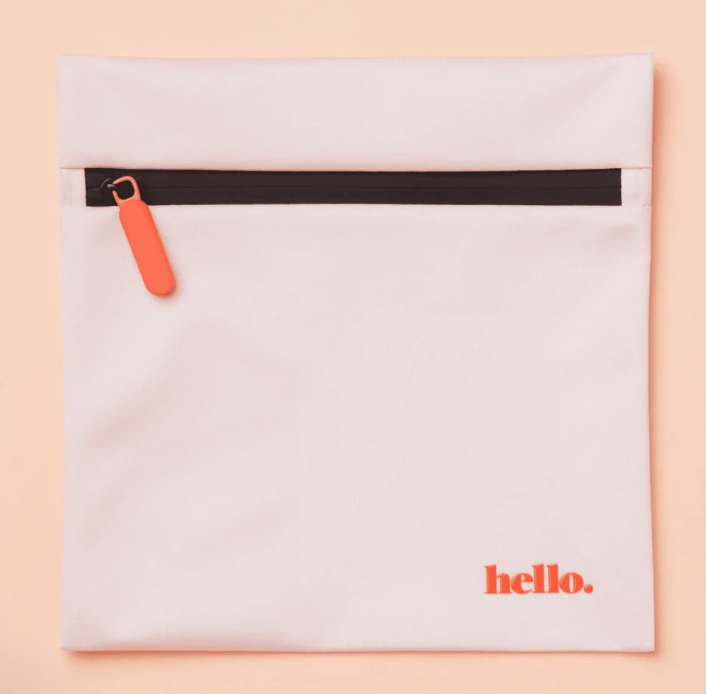 The Hello Wet Bag is a light pink waterproof storage bag with an orange zipper pull and "hello." printed in orange in the bottom right corner, shown on a matching light pink background.